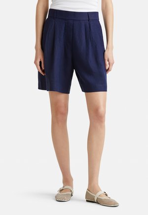 Kobieta w granatowych plisowanych szortach z wysokim stanem i beżowych płaskich butach plecionych, stojąca z rękami swobodnie opuszczonymi wzdłuż ciała.