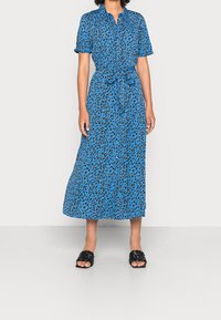 Niebieska sukienka koszulowa z krótkim rękawem w wzór w panterę, zwężana w talii za pomocą związanej wstążki. Noszona z czarnymi plecionymi klapkami.