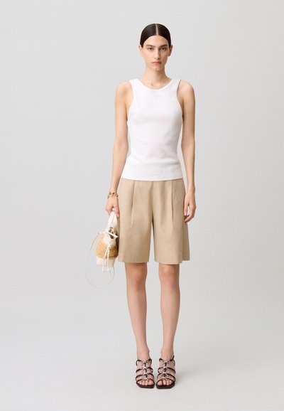 Mujer de pie con una blusa blanca sin mangas, pantalones cortos beige hasta la rodilla, tacones negros con tiras y sosteniendo un pequeño bolso tejido beige.