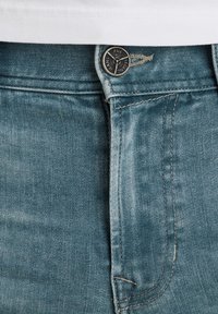 Jeans de mezclilla en azul claro con una textura suave. Presentan un botón de metal con un logo, costura contrastante y pespuntes en la cintura.