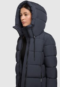 Giacca a vento in blu scuro con un grande cappuccio strutturato, design trapuntato, chiusura frontale con zip e lacci regolabili.