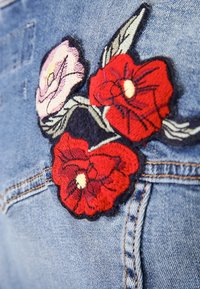 Tkanina dżinsowa o niebieskim odcieniu z haftowanymi kwiatowymi wzorami. Trzy kwiaty w kolorze czerwonym i różowym, z czarnymi konturami i zielonymi liśćmi.