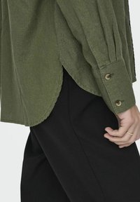 Olivgrünes, langärmliges Shirt mit lockerer Passform, geknöpften Manschetten und genähtem Saum, kombiniert mit schwarzen Hosen.