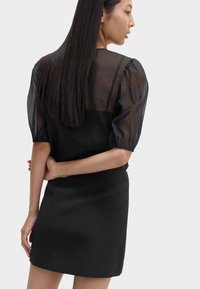 Schwarzes Kleid mit einem durchsichtigen Oberteil mit Puffärmeln und einem taillierten Rock. Glatte Textur; klassisches Design mit modernem Touch. Verborgener Reißverschluss hinten.
