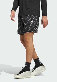 Mustat urheilushortsit, joissa on valkoisia ja harmaita diagonaalisia raidoita, Adidas-logo lahkeessa, yhdistettynä mustiin sukkiin ja ainutlaatuisiin hopeisiin juoksukenkään.