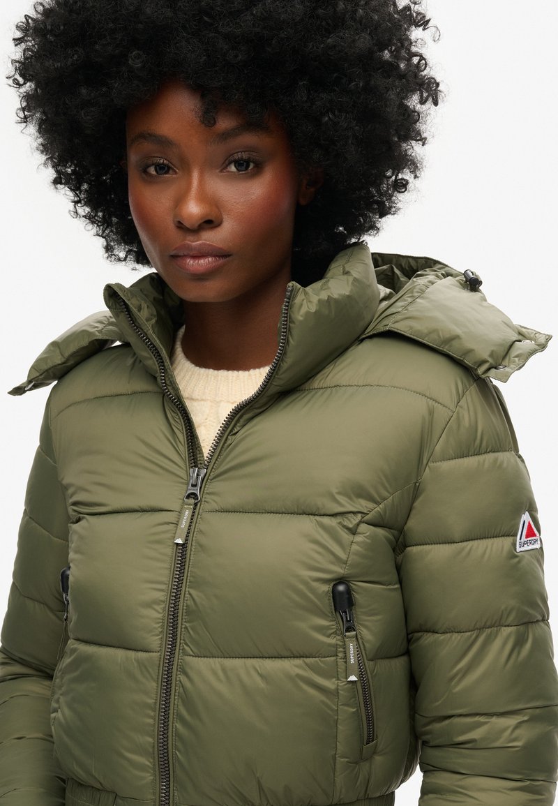 Olijfgroene pufferjack met ritszakken, hoge kraag en afneembare capuchon. Heeft een gequilt uiterlijk en een klein logo op de mouw.