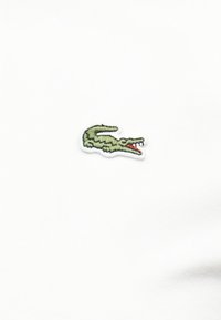 Πράσινο κεντημένο λογότυπο κροκοδείλου με ανοιχτό κόκκινο στόμα σε λευκό ύφασμα.