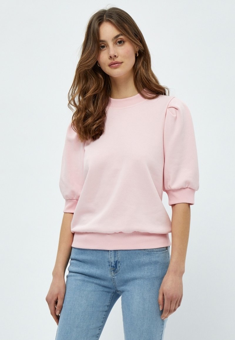 Rosa sweatshirt med puffärmar, ribbad halsringning och fåll. Mjuk tygkvalitet, avslappnad passform, kombinerad med ljusblå denimjeans.