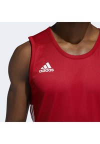 Débardeur rouge en tissu en maille. Présente un col rond et un design sans manches. Logo Adidas blanc sur la poitrine gauche.