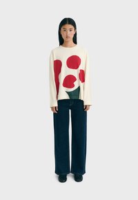 Woman standing against a plain background wearing a cream sweater with red and green abstract shapes, dark wide-leg jeans, and black shoes.