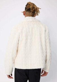 Personne avec des dreadlocks attachées, portant une veste texturée blanche et moelleuse ainsi que des pantalons noirs, vue de dos sur fond uni.