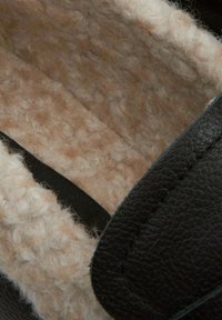 Zwarte lederen slipper met een gestructureerde crèmekleurige wolvoering. Heeft een gladde buitenkant en zacht, donzig binnenmateriaal.