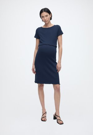 Kobieta w ciąży stojąca w gładkiej granatowej sukience ciążowej z krótkim rękawem, z postrzępionymi krawędziami, i czarnych sandałach na słupku z rzemykami.