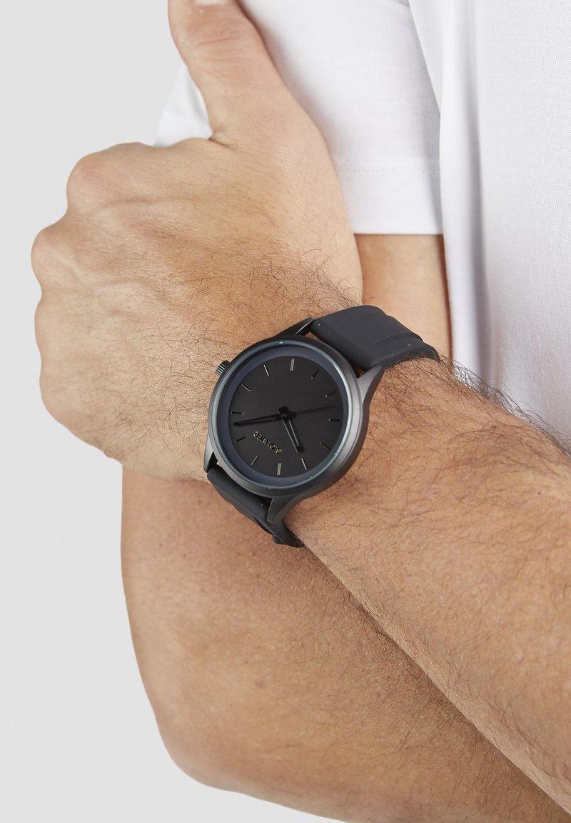 Montre analogique noire à cadran rond au design minimaliste, dotée d'un bracelet en silicone et de marqueurs d'heure argentés.