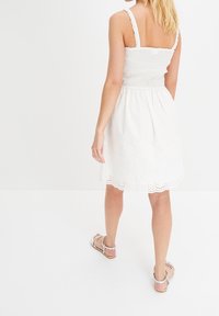 Vit bomulls klänning med smockad bodice och volangärmar, med en scallopad nederkant och ögonmönster nära fållkanten.