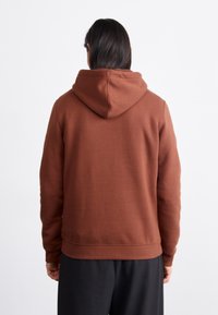 Brown hoodie made of a textured fabric, featuring a hood and cuffed sleeves. The back is smooth with a simple design. Paired with black pants.