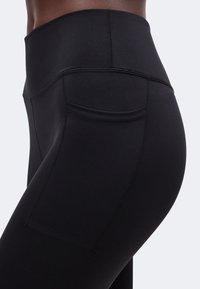 Gros plan d'une personne portant des leggings noirs taille haute avec une poche latérale sur la cuisse, sur un fond uni.