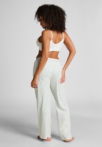 Vrouw met krullend haar, gekleed in een witte crop top en losse witte broek met een blauw bloemenprint, staande op blote voeten tegen een effen achtergrond.