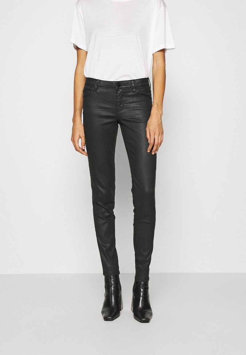 Person som bär svarta slim-fit läderbyxor, svarta klackiga ankelstövlar och en vit kortärmad t-shirt mot en neutral bakgrund.