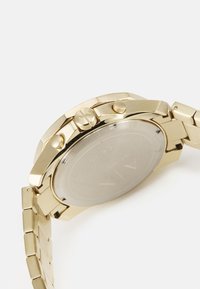 Orologio in metallo dorato con superficie lucida, cassa rotonda, doppio pulsante e bracciale a maglie. Il retro presenta una finitura metallica con il marchio.