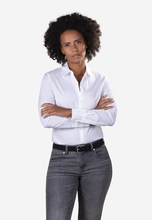 Donna con capelli ricci che indossa una camicia bianca con bottoni e jeans grigi, in piedi con le braccia incrociate davanti a uno sfondo semplice.