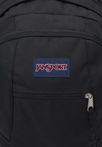 Schwarzer Jansport-Rucksack aus strukturiertem Stoff, mit einer vorderen Reißverschlusstasche und einem blauen Etikett mit dem Markennamen in Rot.