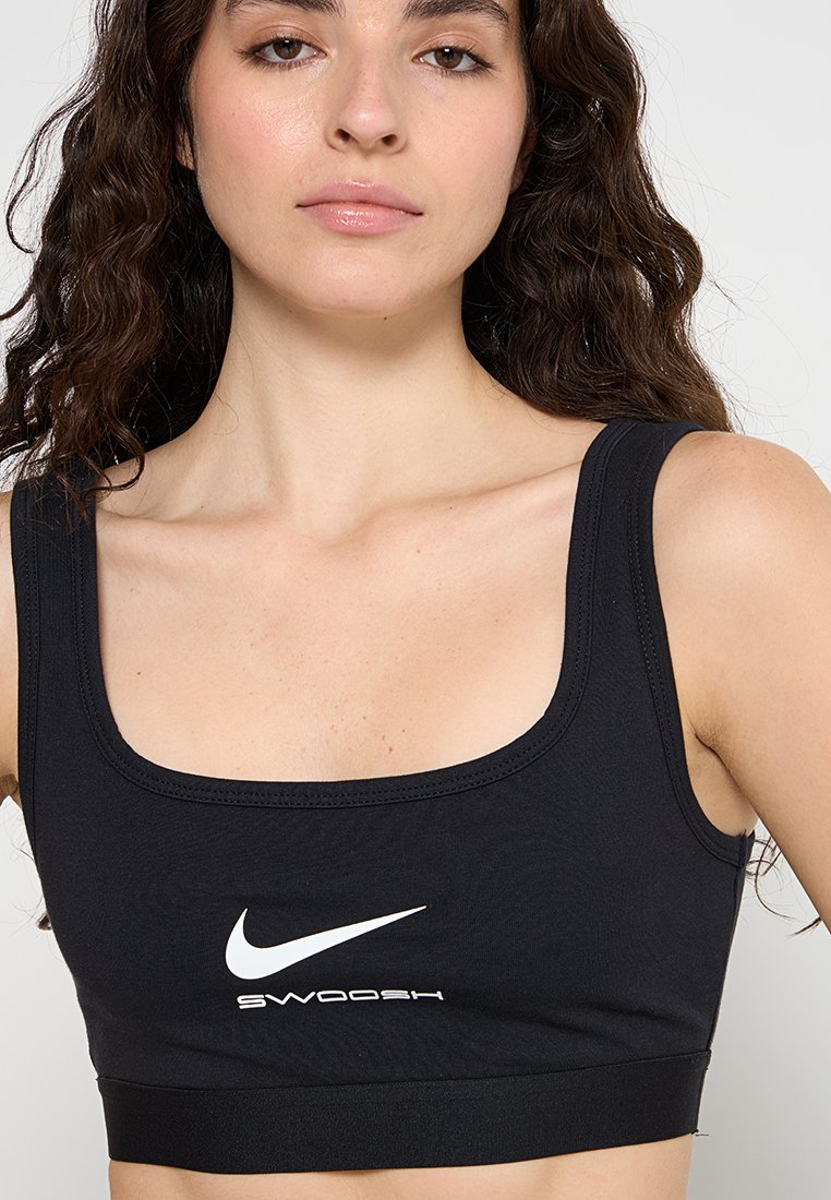 Femme avec de longs cheveux bouclés portant un soutien-gorge de sport Nike noir avec le logo swoosh blanc et le texte "SWOOSH" à l'avant.