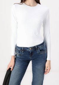 Haut blanc à manches longues en tissu lisse, associé à un jean en denim bleu à coupe classique avec des détails de couture subtils.