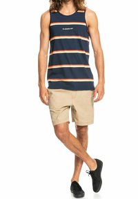 Homme portant un débardeur rayé bleu marine, un short beige et des baskets noires, se tenant avec une jambe croisée sur l'autre.