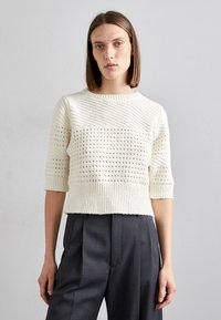 Cremefarbener Rippstrickpullover mit strukturierten horizontalen Streifen und kurzen Ärmeln, kombiniert mit dunkelgrauen maßgeschneiderten Hosen.