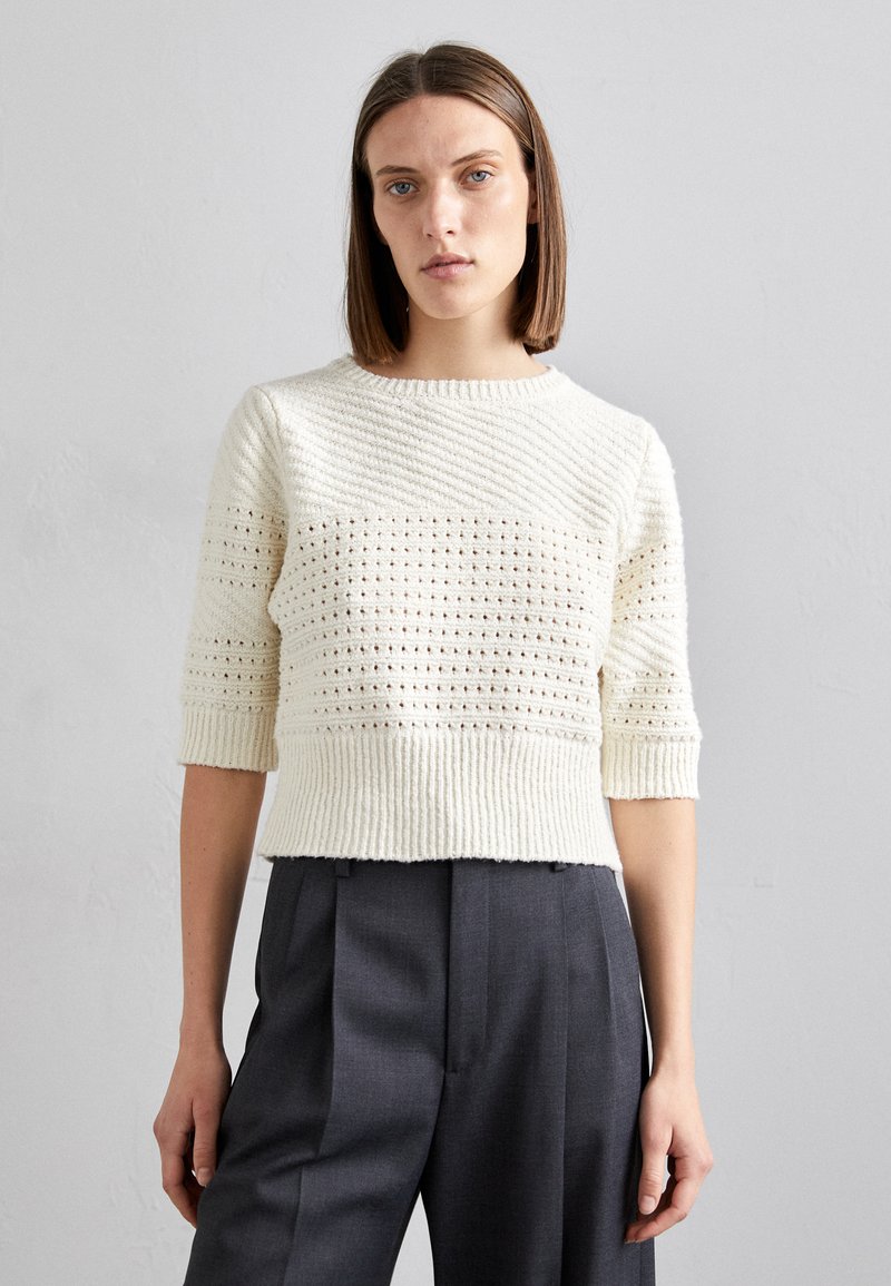 Cremefarbener Rippstrickpullover mit strukturierten horizontalen Streifen und kurzen Ärmeln, kombiniert mit dunkelgrauen maßgeschneiderten Hosen.