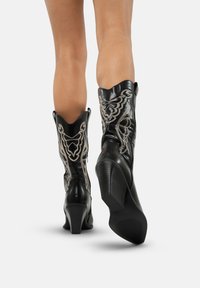 Botas vaqueras de cuero negro con intrincados bordados plateados, punta afilada y tacón apilado. Parte superior redondeada con curvas pronunciadas.