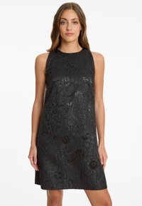 Frau in einem ärmellosen schwarzen Kleid mit einem dezenten Blumenmuster, die vor einem schlichten weißen Hintergrund steht.
