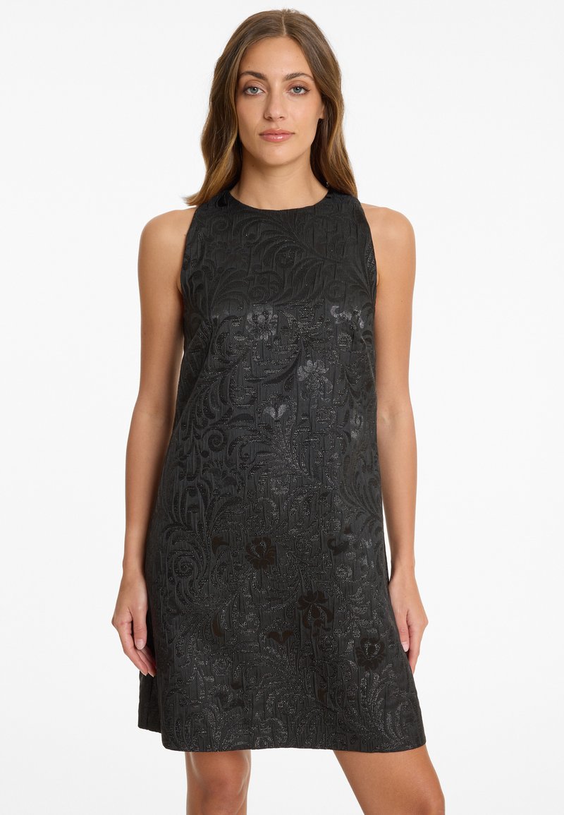 Frau in einem ärmellosen schwarzen Kleid mit einem dezenten Blumenmuster, die vor einem schlichten weißen Hintergrund steht.