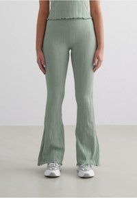 Green ribbed flared pants with a textured surface, featuring a fitted waist and ruffled hem. Worn with grey athletic shoes.