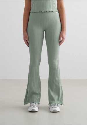 Green ribbed flared pants with a textured surface, featuring a fitted waist and ruffled hem. Worn with grey athletic shoes.