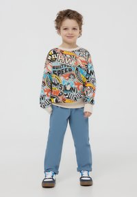 Felpa colorata con grafiche e testo su uno sfondo crema; abbinata a pantaloni azzurri chiari e sneakers blu e bianche.