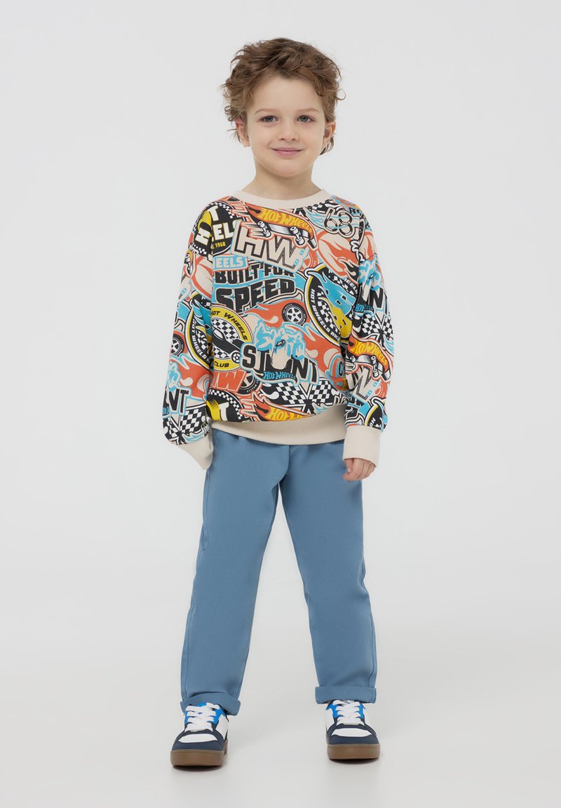 Felpa colorata con grafiche e testo su uno sfondo crema; abbinata a pantaloni azzurri chiari e sneakers blu e bianche.