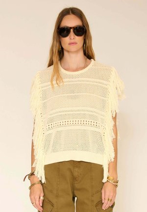 Femme portant de grandes lunettes de soleil sombres, un haut crème en crochet avec des franges, et un pantalon marron, debout devant un fond beige uni.
