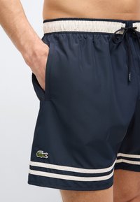 Άνδρας που φοράει μπλε ναυτικό μαγιό με δύο μπεζ ρίγες κοντά στο τελείωμα, ελαστική ζώνη μέσης, κορδόνι και πράσινο πατς με λογότυπο κροκόδειλου.