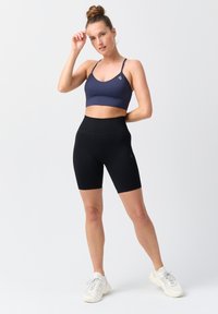 Navy gerippter Sport-BH mit dünnen Trägern, kombiniert mit hochgeschnittenen schwarzen gerippten Radlerhosen und getragen mit weißen Sportschuhen.