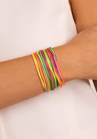 Arm met meerdere dunne elastische armbandjes in felle neonkleuren waaronder roze, geel, groen, oranje en blauw tegen een witte achtergrond.