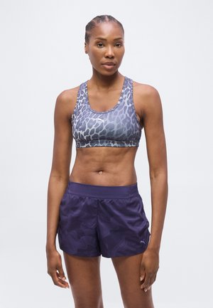 Femme athlétique debout portant un soutien-gorge de sport à motif et un short de sport violet foncé sur un fond clair uni.