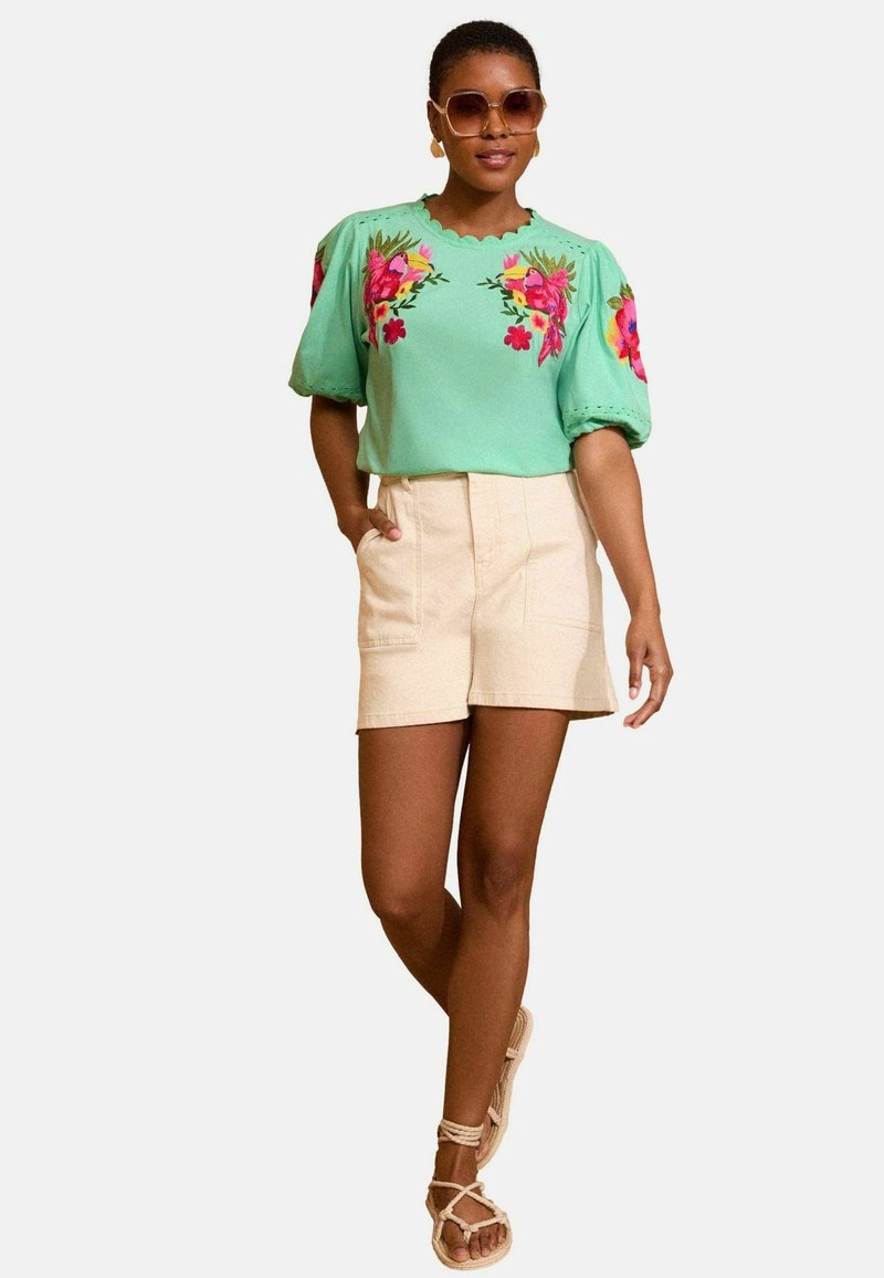 Zöld hímzett blúz puffos ujjakkal, bézs sorttal kombinálva. A modellen barna, pántos szandál és oversized napszemüveg van.