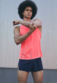 Helles pinkes ärmelloses Sportoberteil mit Netzstruktur, kombiniert mit marineblauen Shorts. Das Modell übt Druck auf den Arm aus, um eine athletische Figur zu präsentieren.