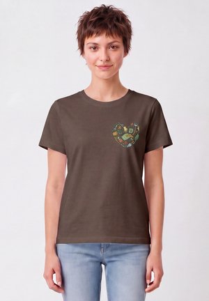 Frau mit kurzen braunen Haaren, die ein dunkelbraunes T-Shirt mit einem kleinen bunten Herz aus Gartengeräten auf der linken Brust trägt, kombiniert mit hellblauen Jeans.