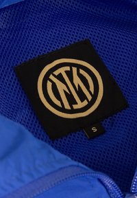 Blauwe stoflabel heeft een zwart patch met een gouden cirkelvormig logo en "NYK" lettering, bevestigd aan een gaasvoering met een maatlabel.