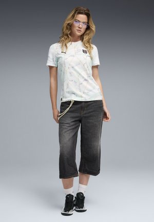 Femme aux cheveux blonds ondulés portant des lunettes, un t-shirt blanc à motifs Puma, un pantalon noir court avec une chaîne de perles, des chaussettes blanches et des baskets noires.