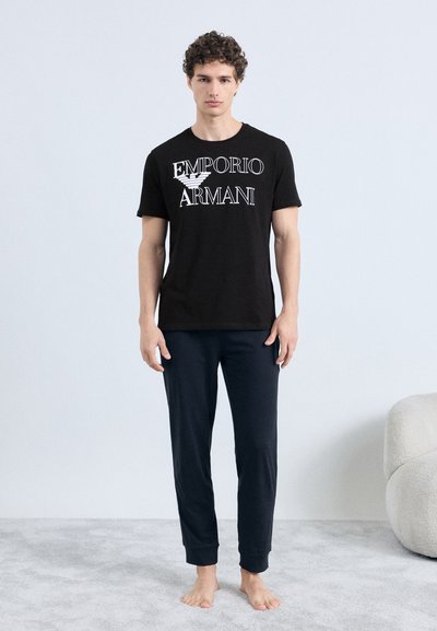 Čierne bavlnené tričko s bielym logom "Emporio Armani", doplnené tmavomodrými bavlnenými nohavicami. Model stojí bosý na svetlom koberci.