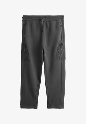 Junger Mann steht, trägt hellgraues T-Shirt, dunkelgraue Jogginghose, graue Sneakers und einen leichten Pullover über den Schultern.